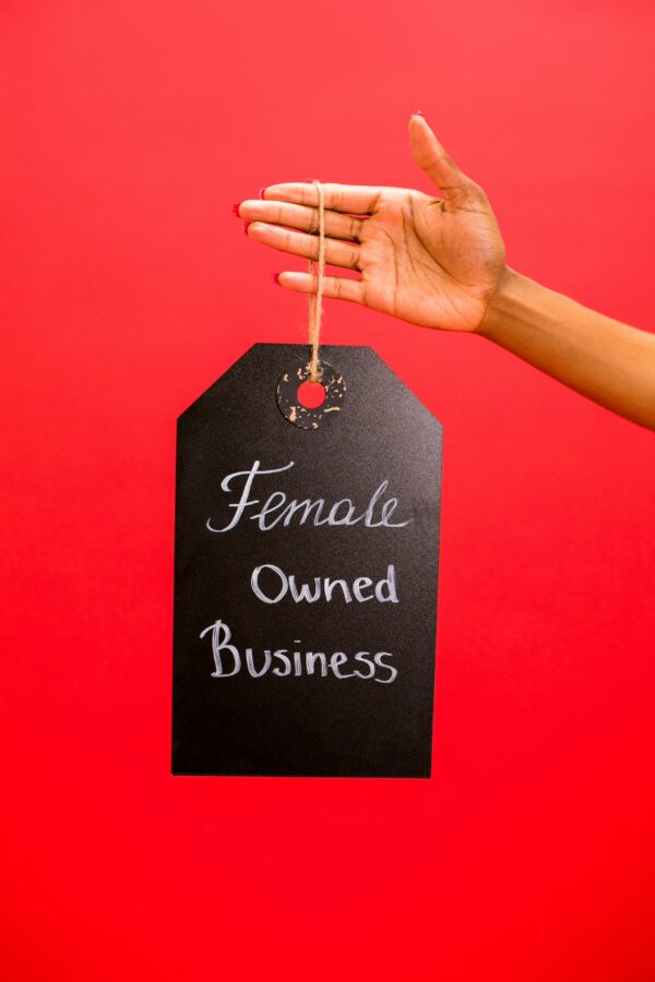 A hand holding a tag sign reading 'Female Owned Business' against a vivid red background.